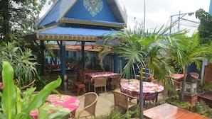 Ein Terrassenbereich im Freien mit Tischen und Stühlen, umgeben von Blumentöpfen und einer Struktur mit blauem Dach.