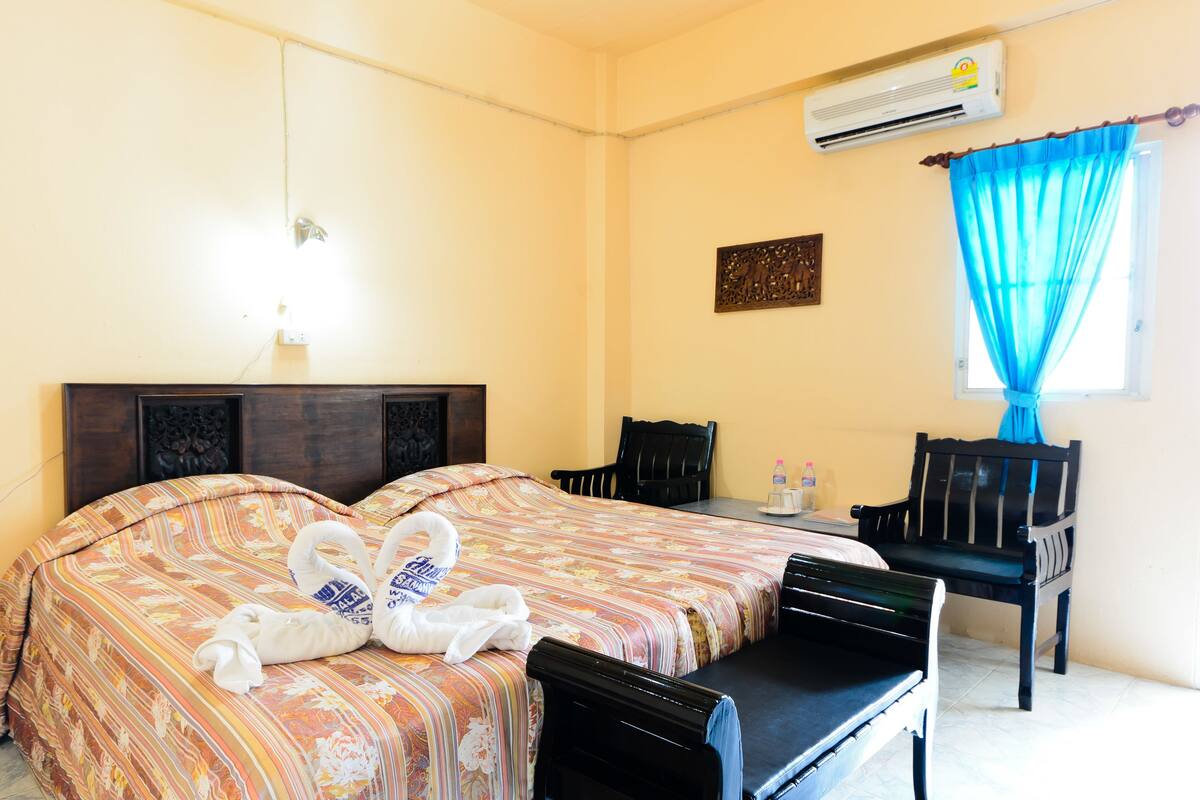 A hotel room with a bed, two chairs, a table, an air conditioning unit, and a window with blue curtains.