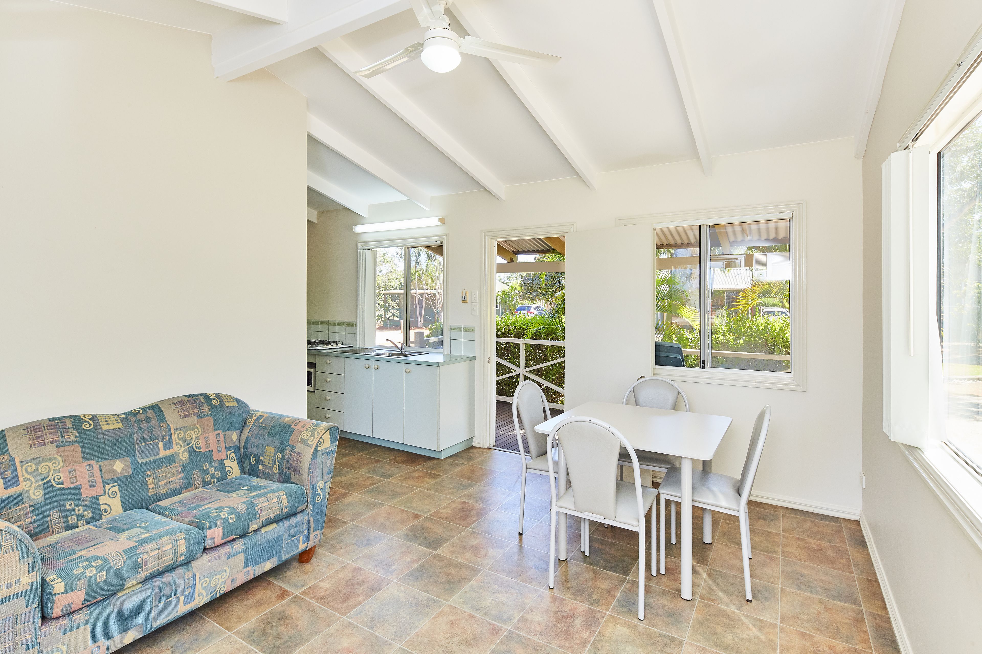 A spacious living area with a sofa, dining table, and chairs. There is a ceiling fan, a window with a view of greenery, and a kitchen area in the background.