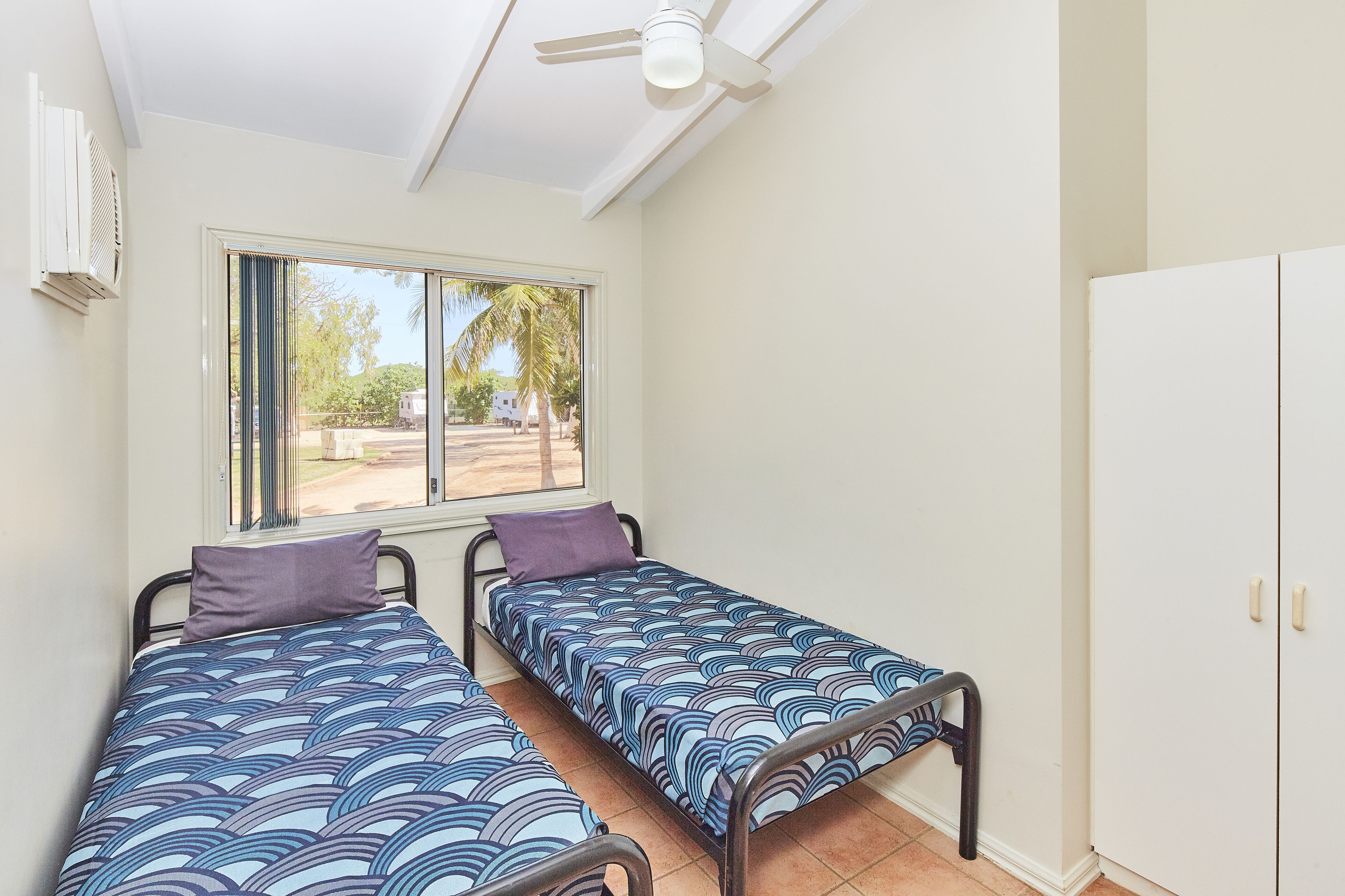 Una habitació amb dos llits individuals, una finestra amb cortines, un ventilador de sostre i un armari.