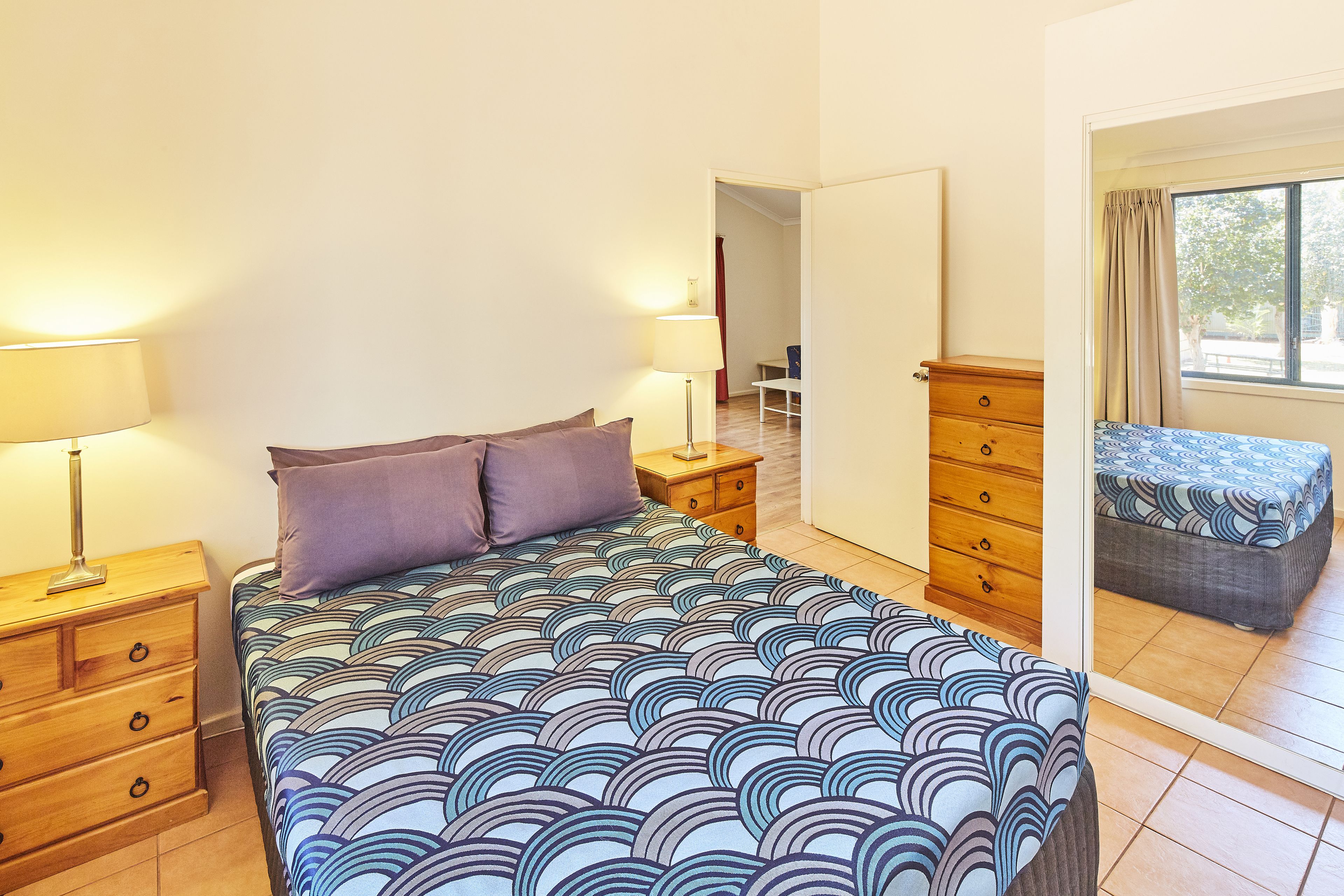 Quarto com cama, duas mesas de cabeceira, cômoda e janela com cortinas.