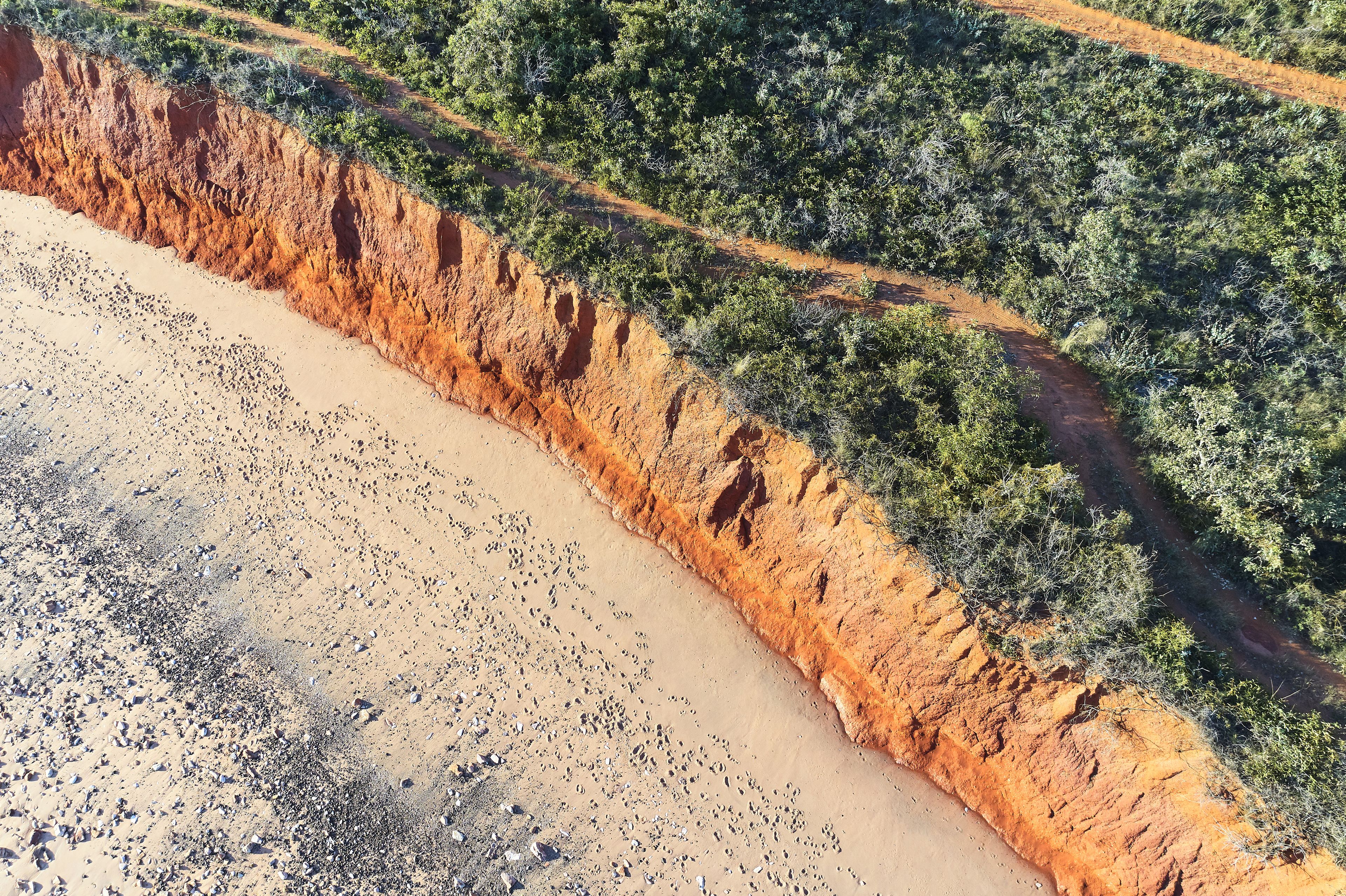 Vista aérea de una zona costera con playa de arena, un terraplén de tierra roja y vegetación verde densa.
