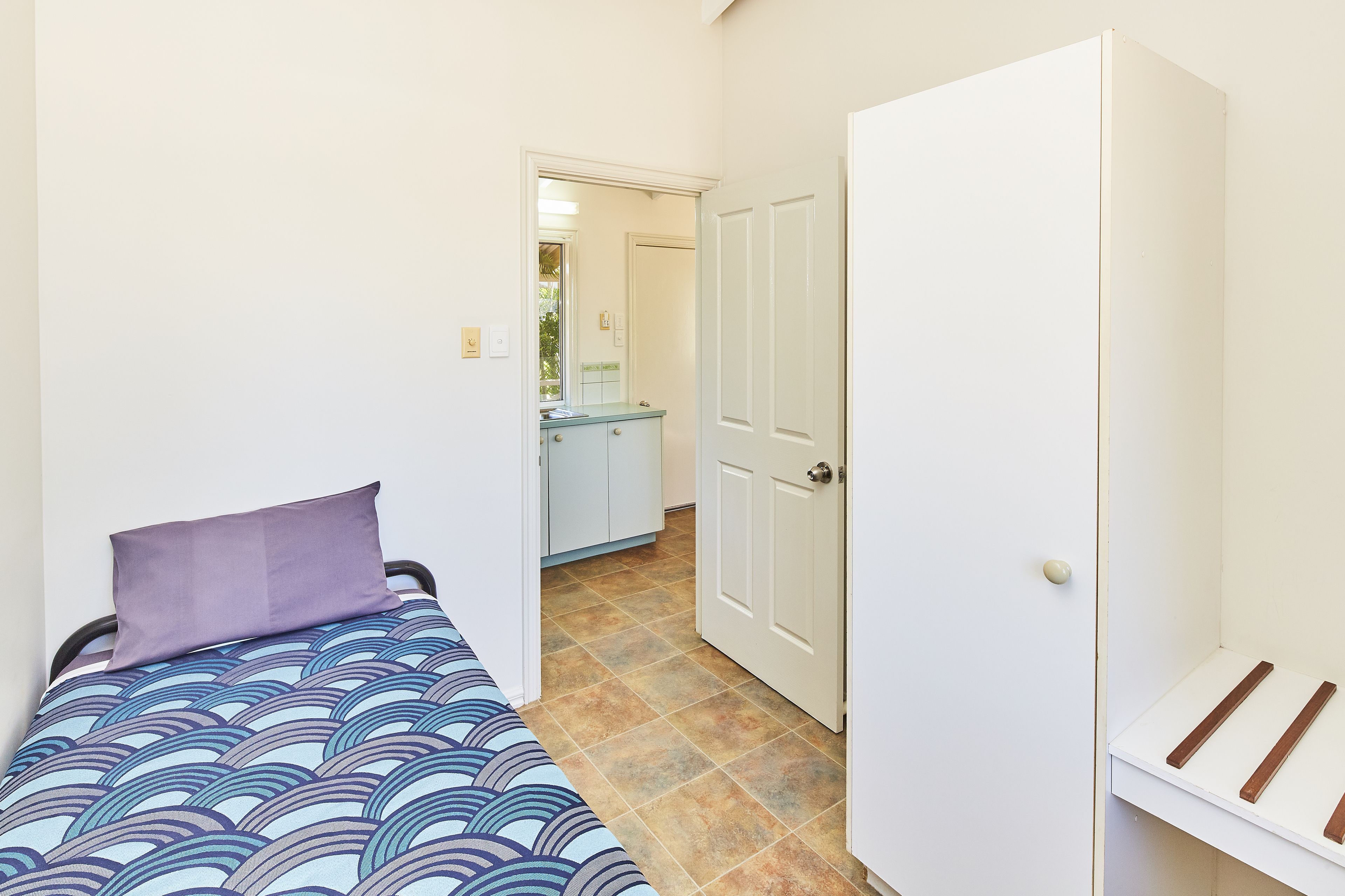 A small, narrow room with a bed, a wardrobe, and a door leading to a bathroom.