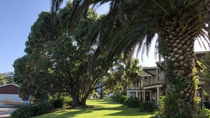 A well-maintained lawn with a large tree and a palm tree, a building with a balcony, and a clear sky. - Half Moon Bay Motel (Auckland)