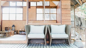 A wicker sofa with white cushions on a wooden deck, large windows, and a rustic interior. - Koh Munnork Private Island (Koh Mun Nork)