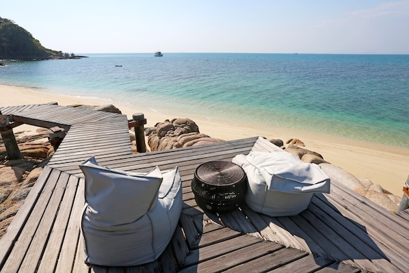 Eine Holzterrasse mit weißen Kissen und einem gewebten Korb, mit Blick auf einen Strand mit klarem türkisblauem Wasser und einem entfernten Boot.