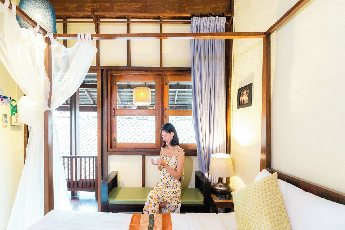 A room with a bed, a window with curtains, a bedside table with a lamp, and a chair.