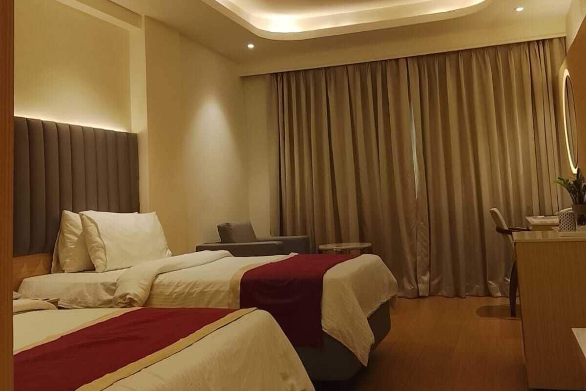 Habitación de hotel con dos camas, un escritorio, una silla, un ventilador de techo y un ventanal grande con cortinas.