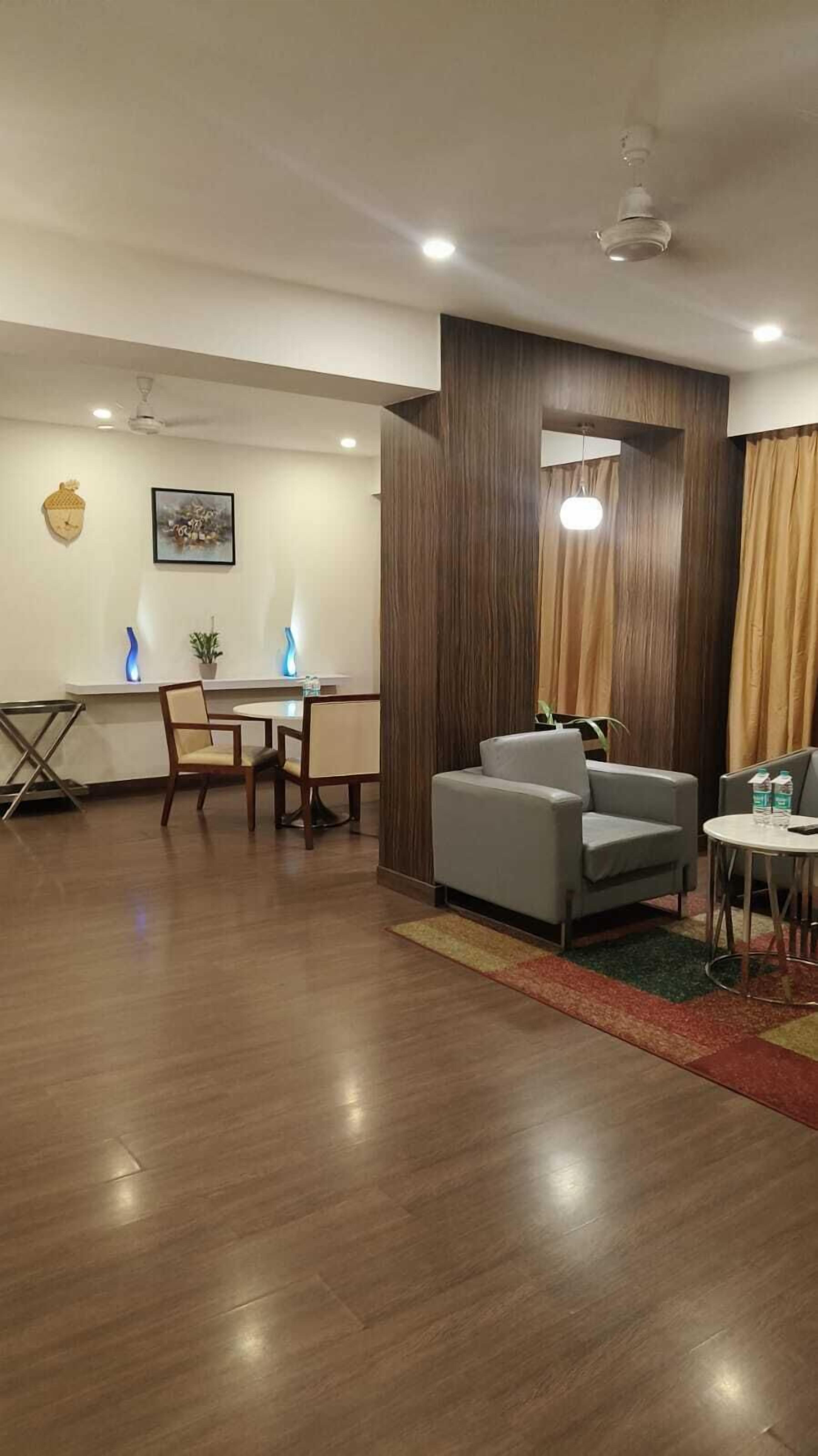 A modern hotel room with a wooden floor, a dining area with a table and chairs, a sofa, and a small table with a lamp.