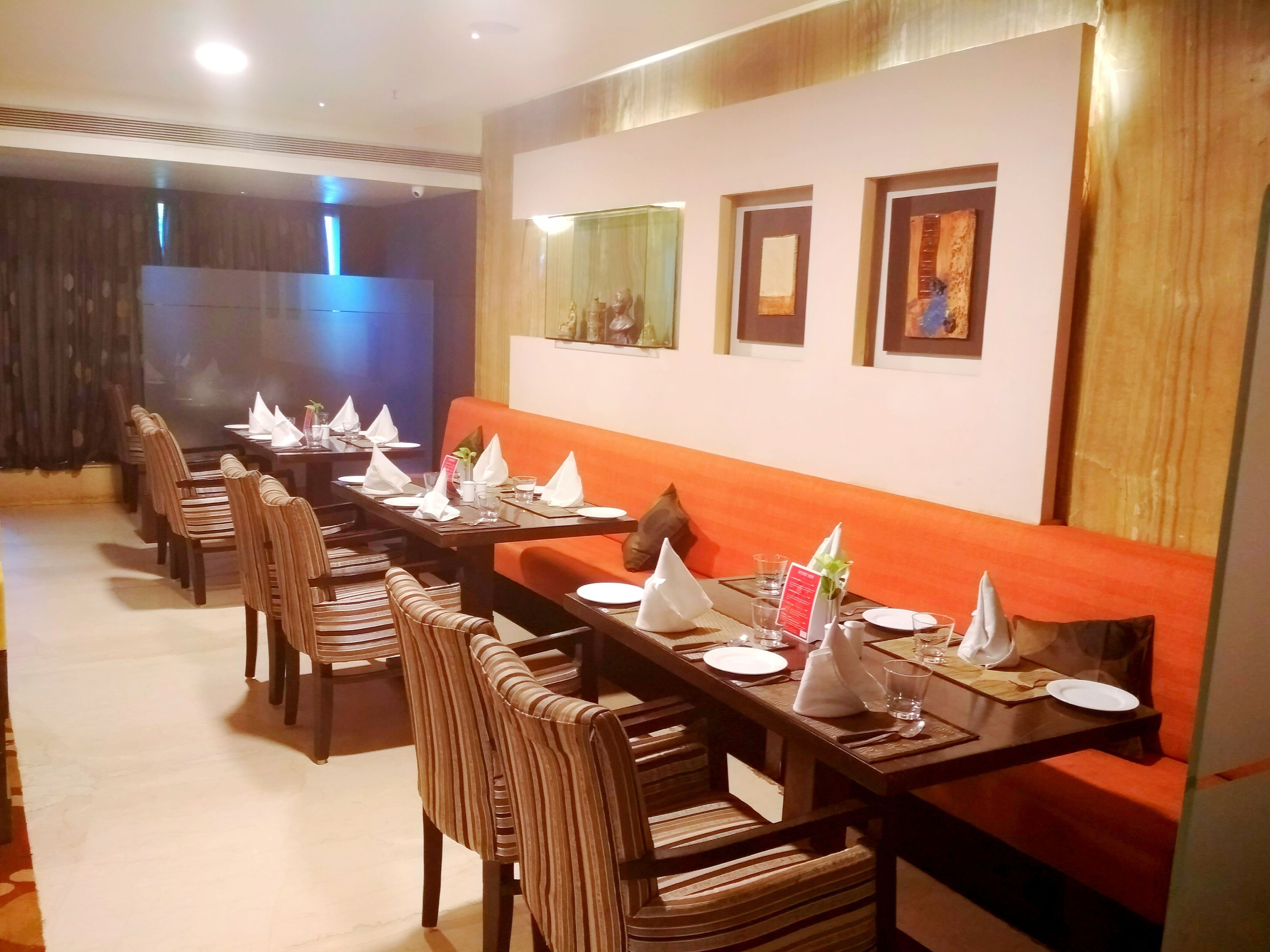 A restaurant with long tables and striped chairs, a red cushioned bench, and framed artwork on the wall.