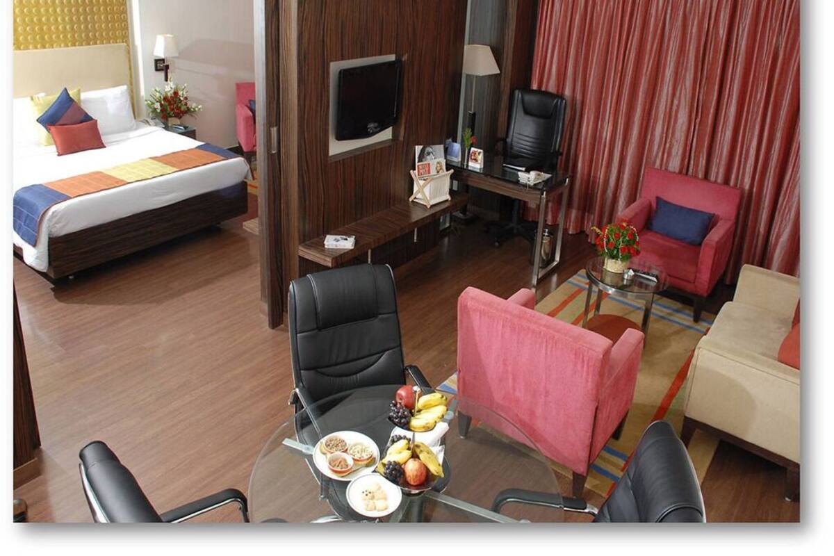 Habitación de hotel con cama, escritorio con computadora, televisor, mesa de comedor con fruta y dos sillas.