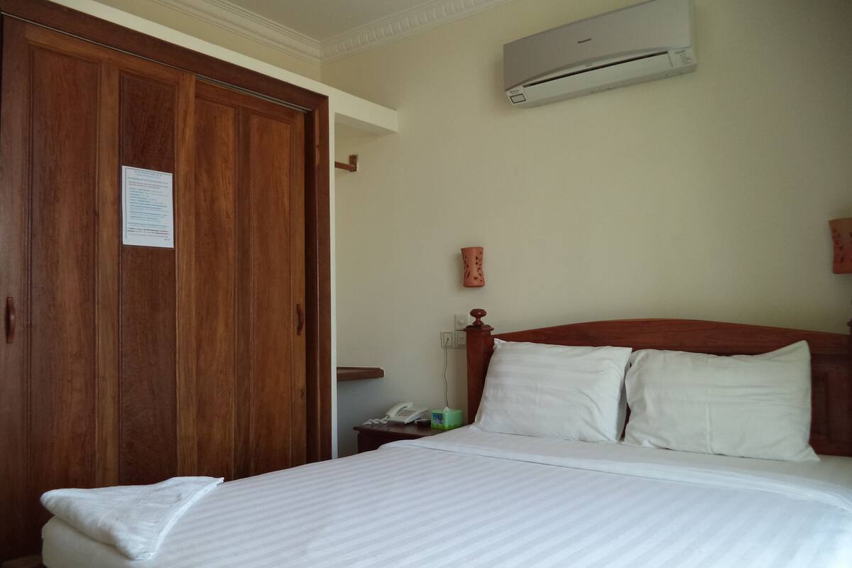 Ein Hotelzimmer mit einem Holzschrank, einem Bett mit weißer Bettwäsche, einem Nachttisch, einem Telefon und einer Klimaanlage an der Wand.