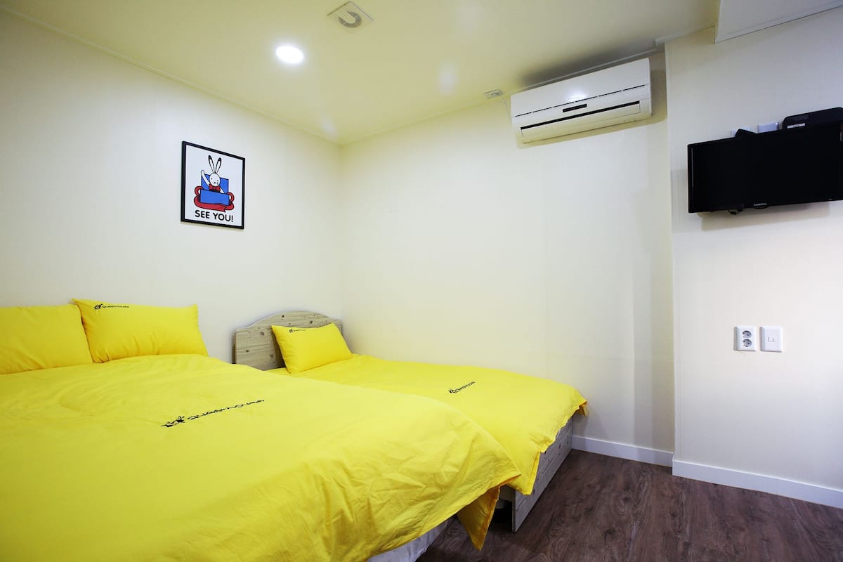 A room with two beds, each with a yellow bedspread and matching pillows, a mounted air conditioner, a flat-screen TV, and a framed picture on the wall.