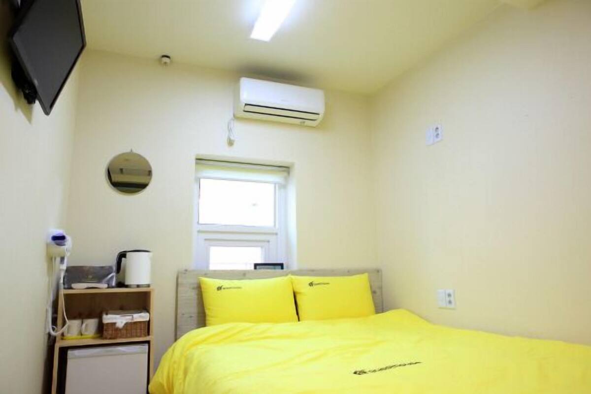 A small, bright room with a bed, a shelf with a kettle, and an air conditioner.