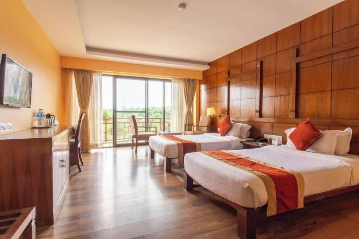 A hotel room with two beds, a desk, a chair, a TV, and a balcony with a view.