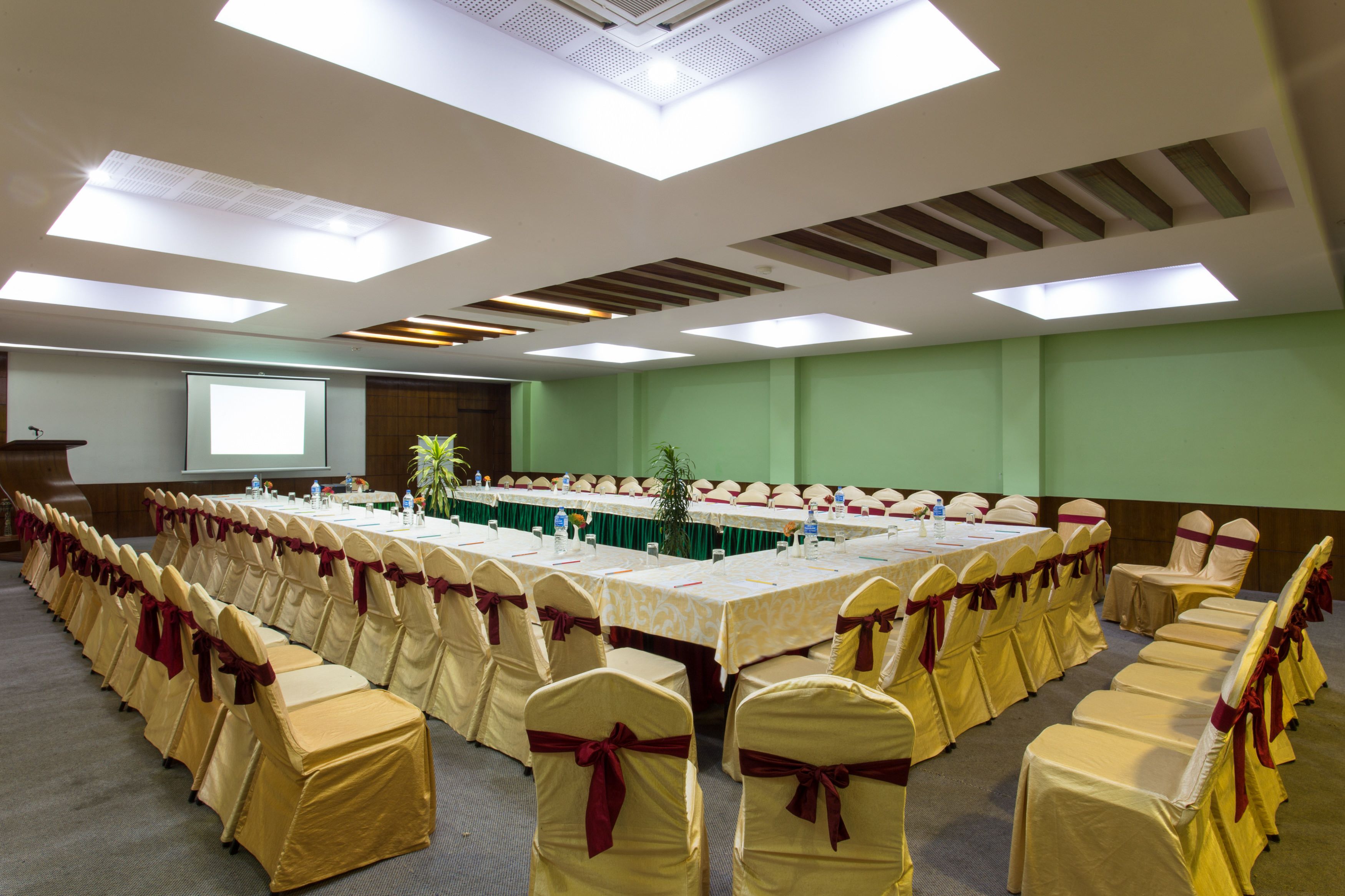 Un salón de conferencias con una larga mesa, sillas con cintas rojas y una pantalla de proyector.