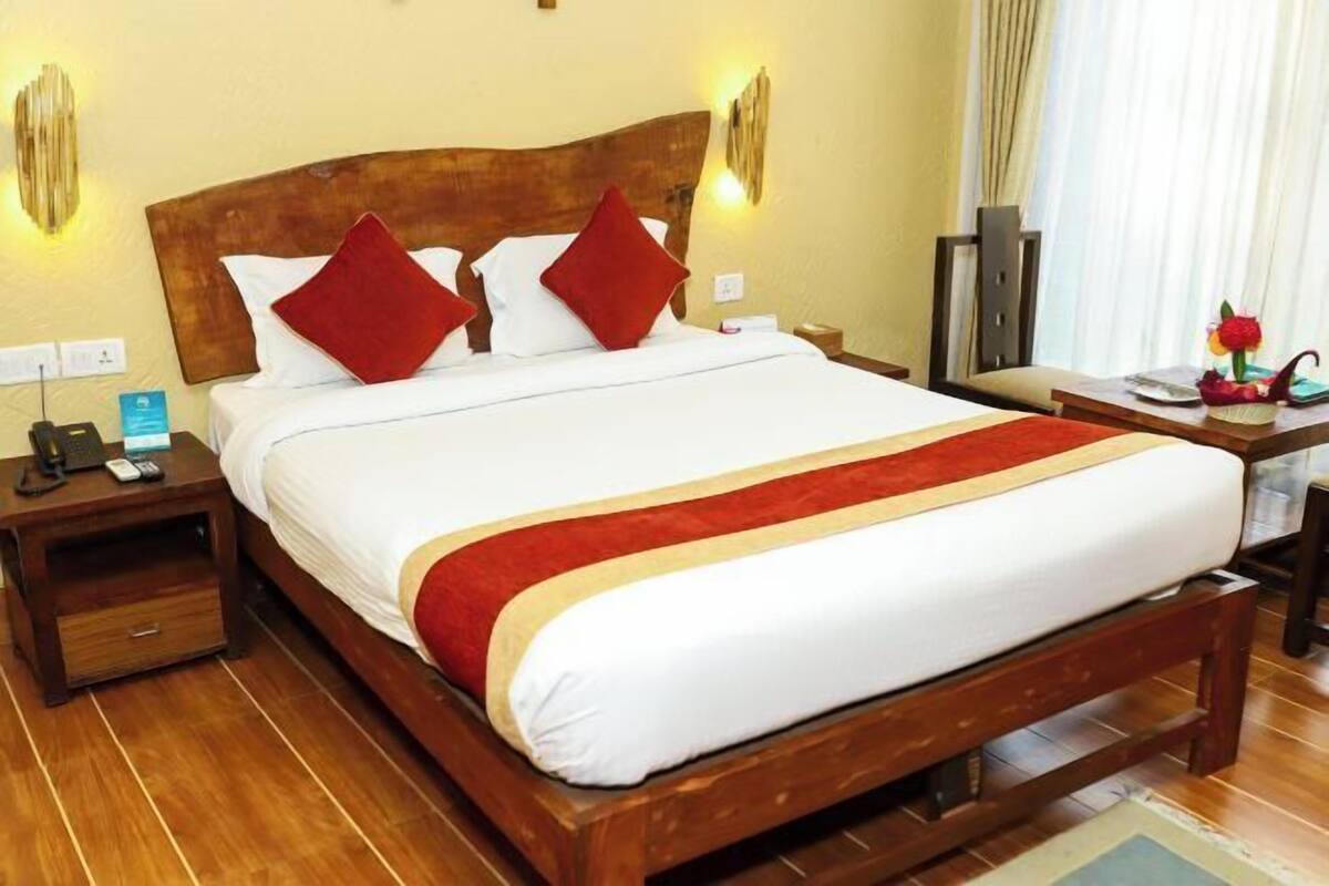 A hotel room with a wooden bed, white bedding, red pillows, a wooden nightstand, a bedside table, and a lamp.