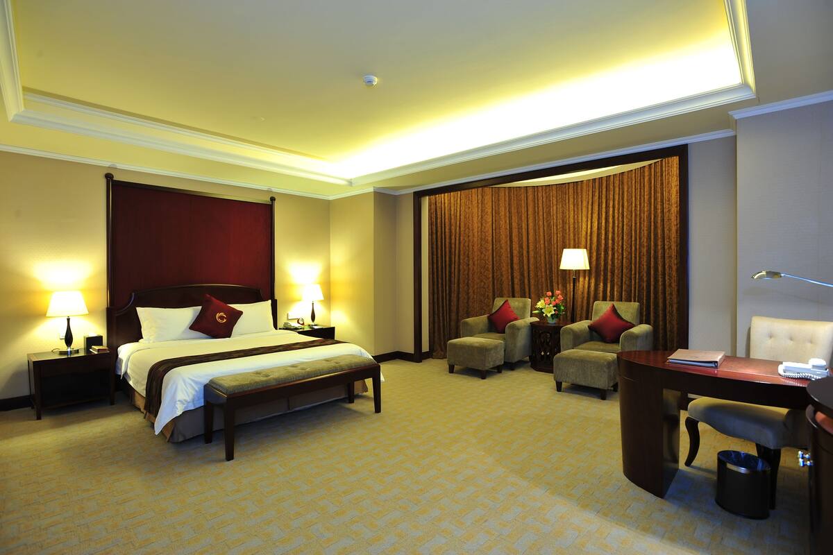 Ein Hotelzimmer mit einem großen Bett, zwei Nachttischen mit Lampen, einem Schreibtisch mit Stuhl und einem großen Fenster mit Vorhängen.
