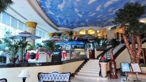 Eine Hotelhalle mit einer imposanten Treppe, einer Deckenmalerei und einem Empfangsbereich mit Palmen und Sonnenschirmen.