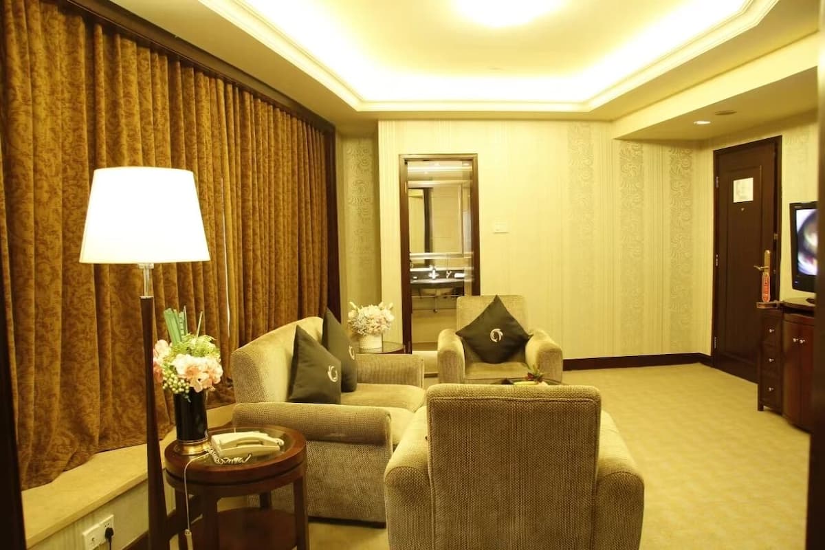 Ein Hotelzimmer mit einer Sitzecke, ausgestattet mit zwei Sesseln und einem Sofa, einem kleinen Beistelltisch mit Lampe und Blumen, einem Fernseher auf einem Holzschrank und einem Spiegel an der Wand.