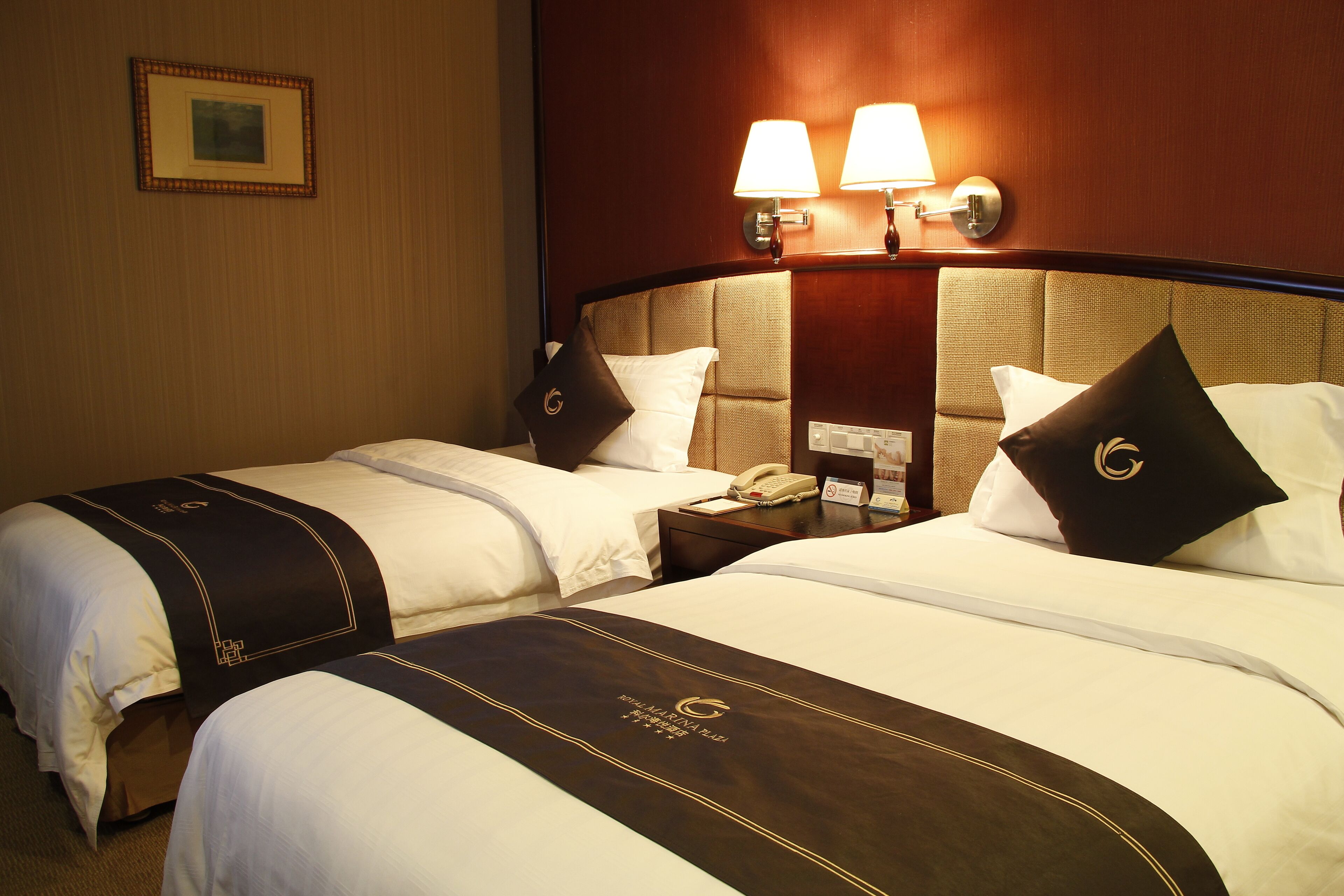 A hotel room with two beds, a bedside table with a phone, and a wall-mounted lamp.