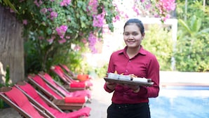 A person holding a tray with food items, standing in front of a pool with red lounge chairs and lush greenery. - SiZen Retreat & Spa (Siem Reap)