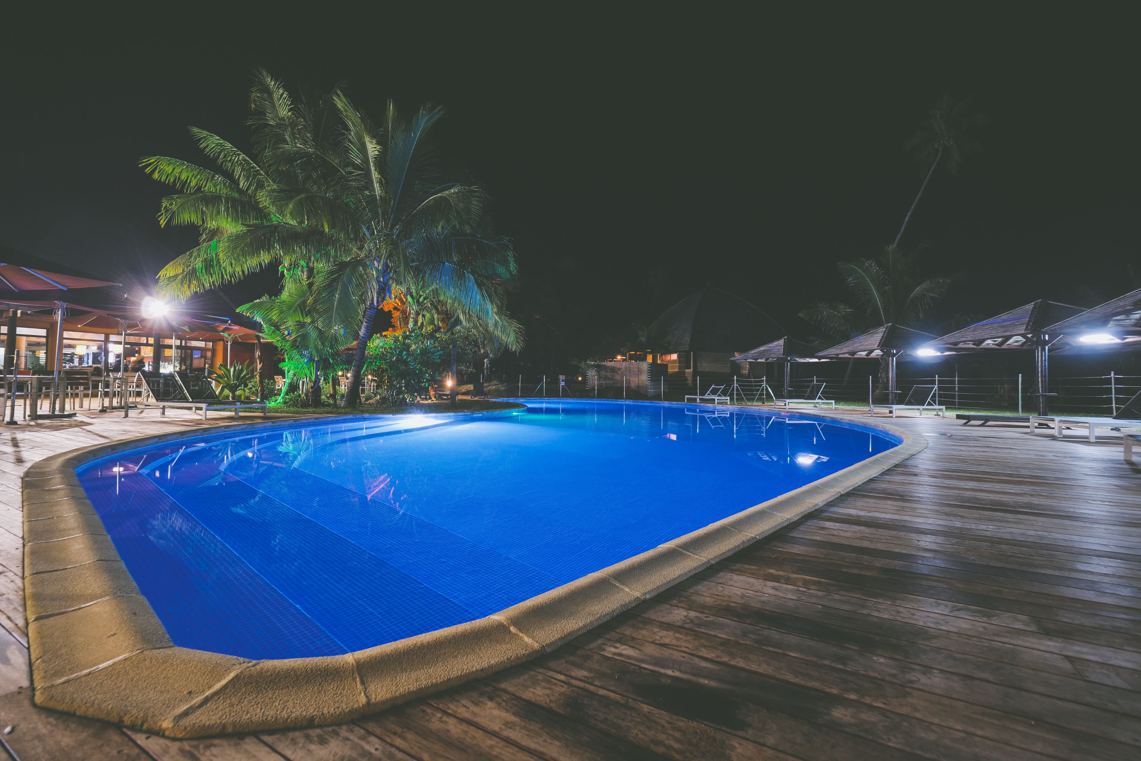 Uma piscina com deck de madeira, rodeada por palmeiras e áreas de estar ao ar livre.