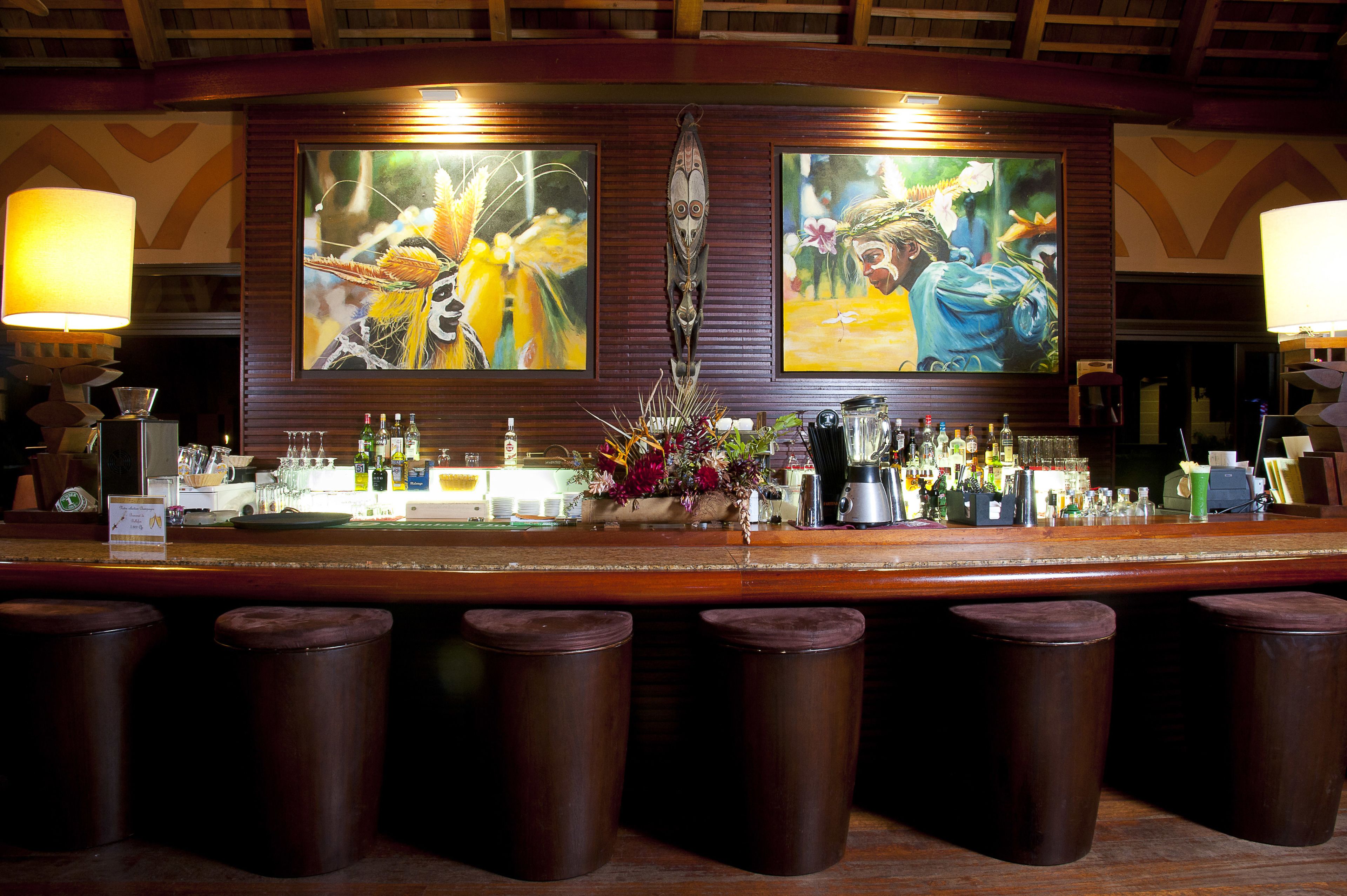 A bar area with a wooden counter, two large paintings, a vase of flowers, and several stools.