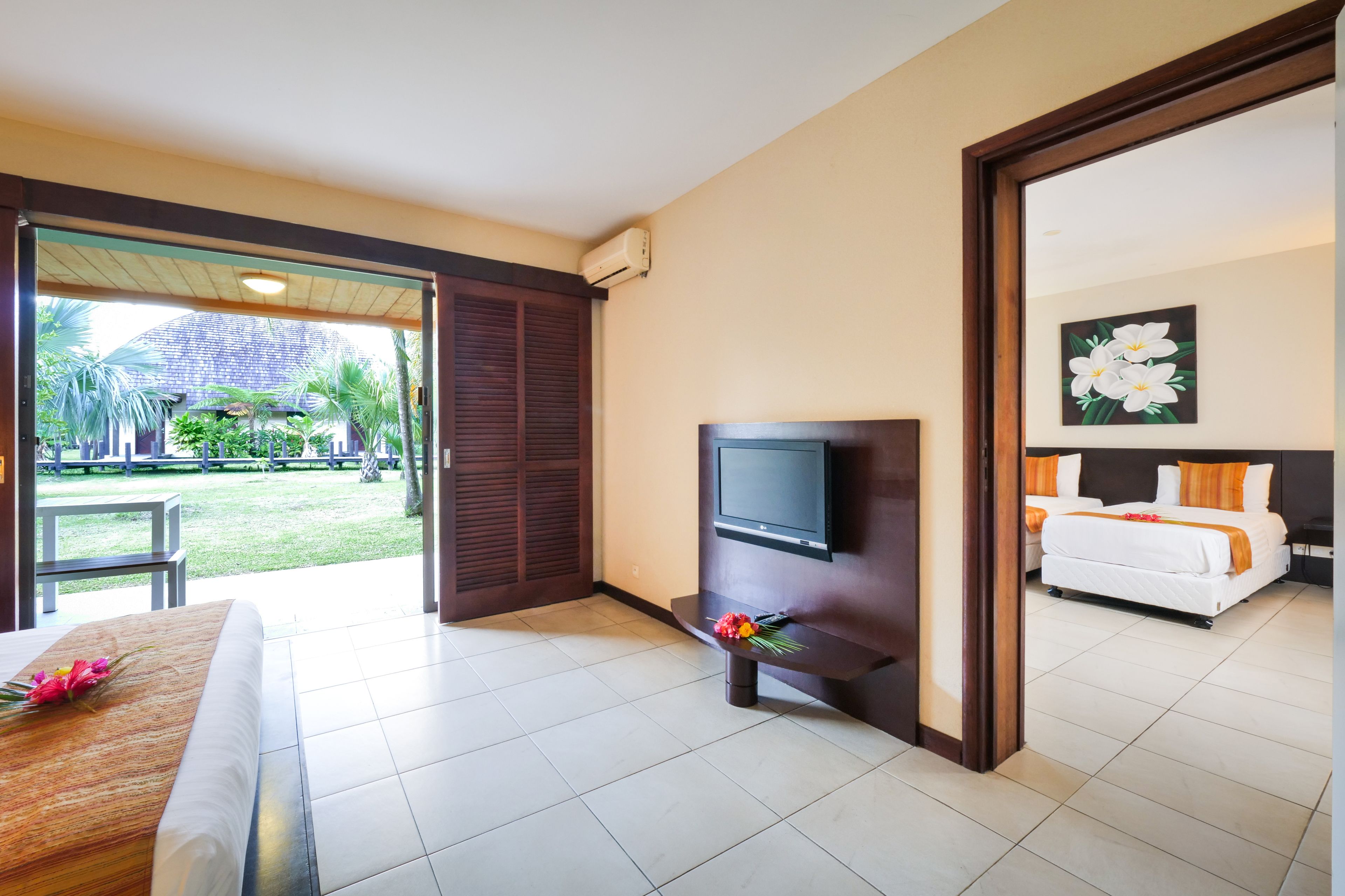 A hotel room with a bed, a TV, a small table with a flower, and a view of the garden and pool area.