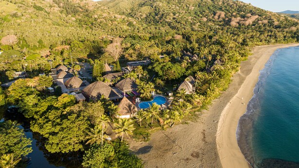 Eine Vogelperspektive zeigt einen Urlaubsort mit mehreren Bungalows, einem Swimmingpool und einem Strand.