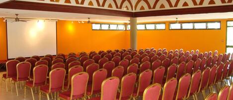 Un salón de conferencias con filas de sillas rojas y una pantalla de proyección blanca.