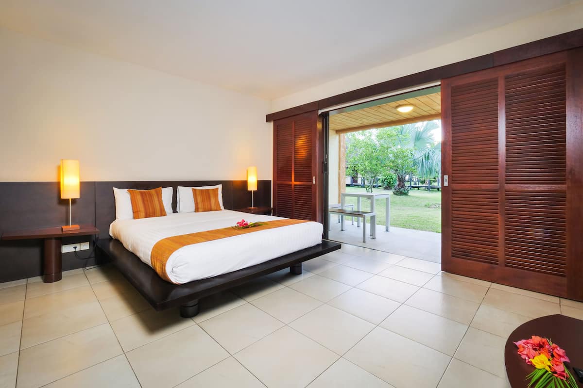 Quarto de hotel com uma cama grande, duas luminárias de cabeceira, um painel de cabeceira de madeira e vista para um jardim por uma porta de correr.