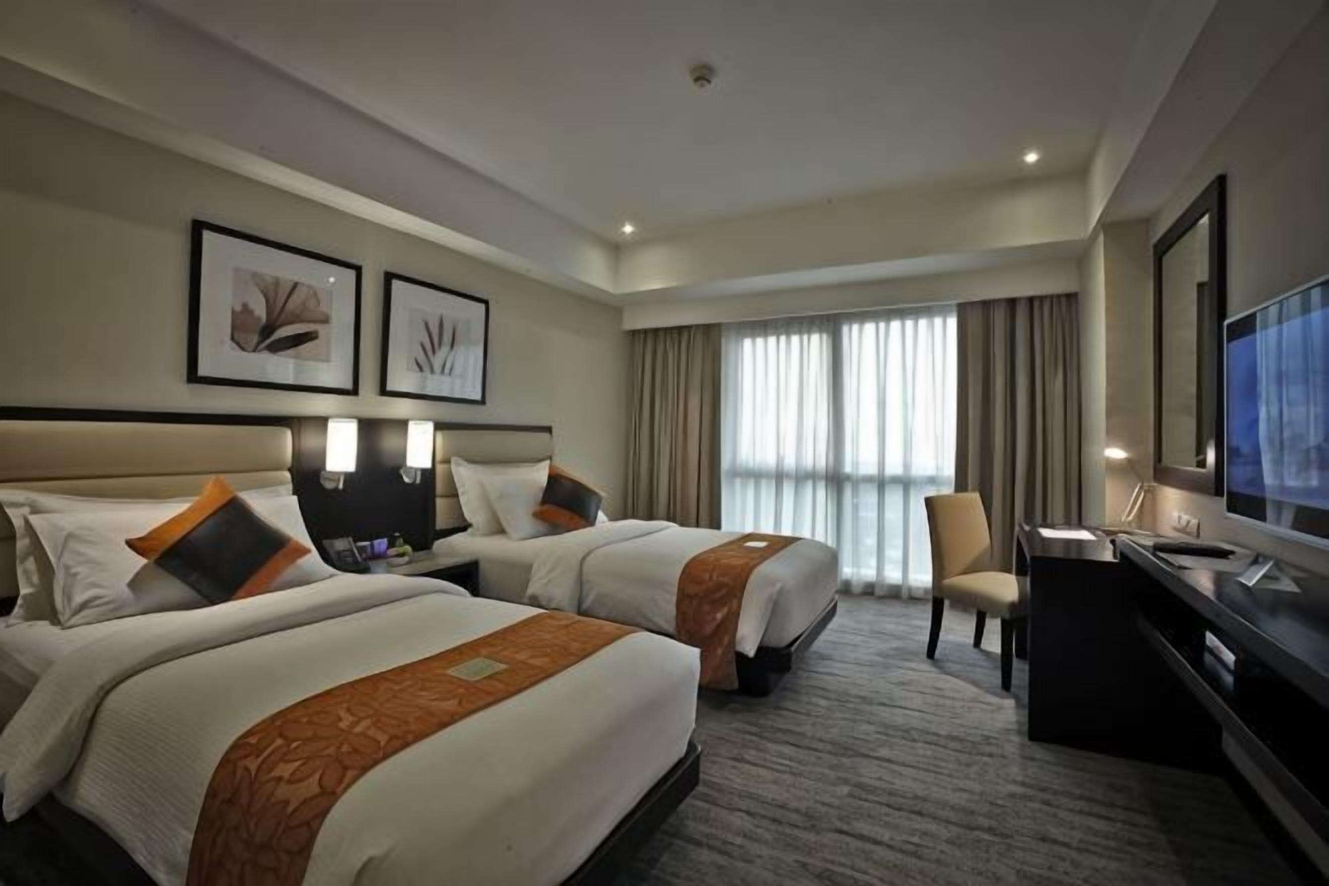 A hotel room with two beds, a desk, a chair, a TV, and framed artwork on the wall.