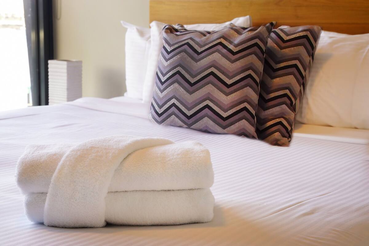 Una cama bien tendida con sábanas blancas, dos almohadones decorativos con estampado de rombos y una toalla blanca doblada sobre la cama.