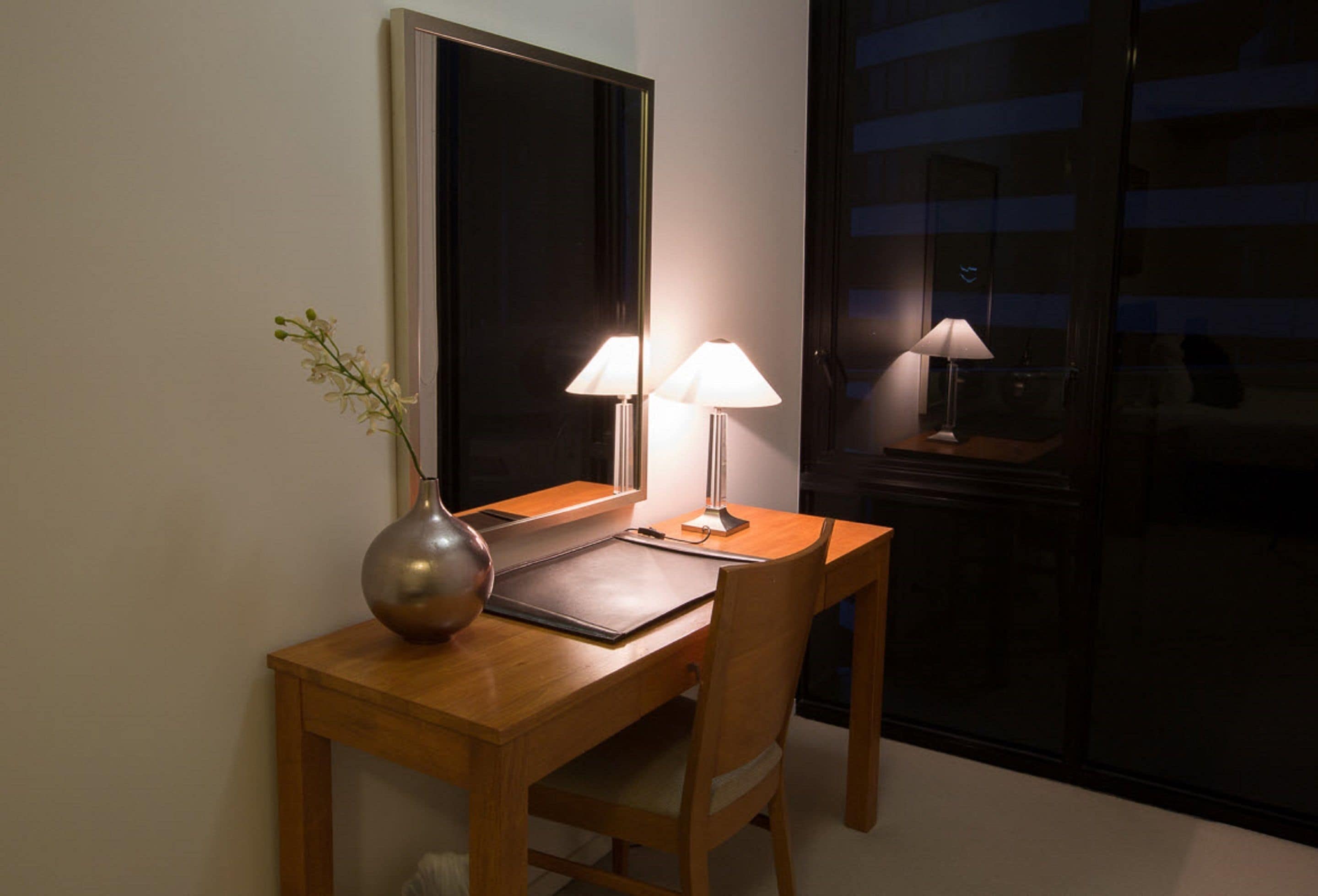 A wooden desk with two lamps, a vase with flowers, and a mirror.