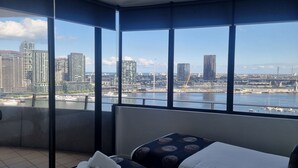 A hotel room with a large window offering a city view, a bed with a patterned bedspread, and a stack of towels on the floor.