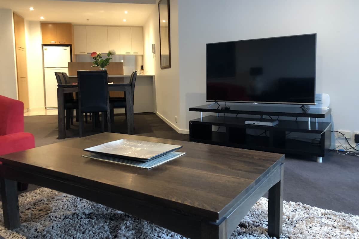 Una sala de estar moderna con un televisor de pantalla plana, un comedor con sillas y una mesa, y una cocina al fondo.