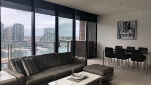 A modern living room with a gray sectional sofa, a white coffee table, and a dining area with black chairs. Large windows offer a view of a cityscape and marina.