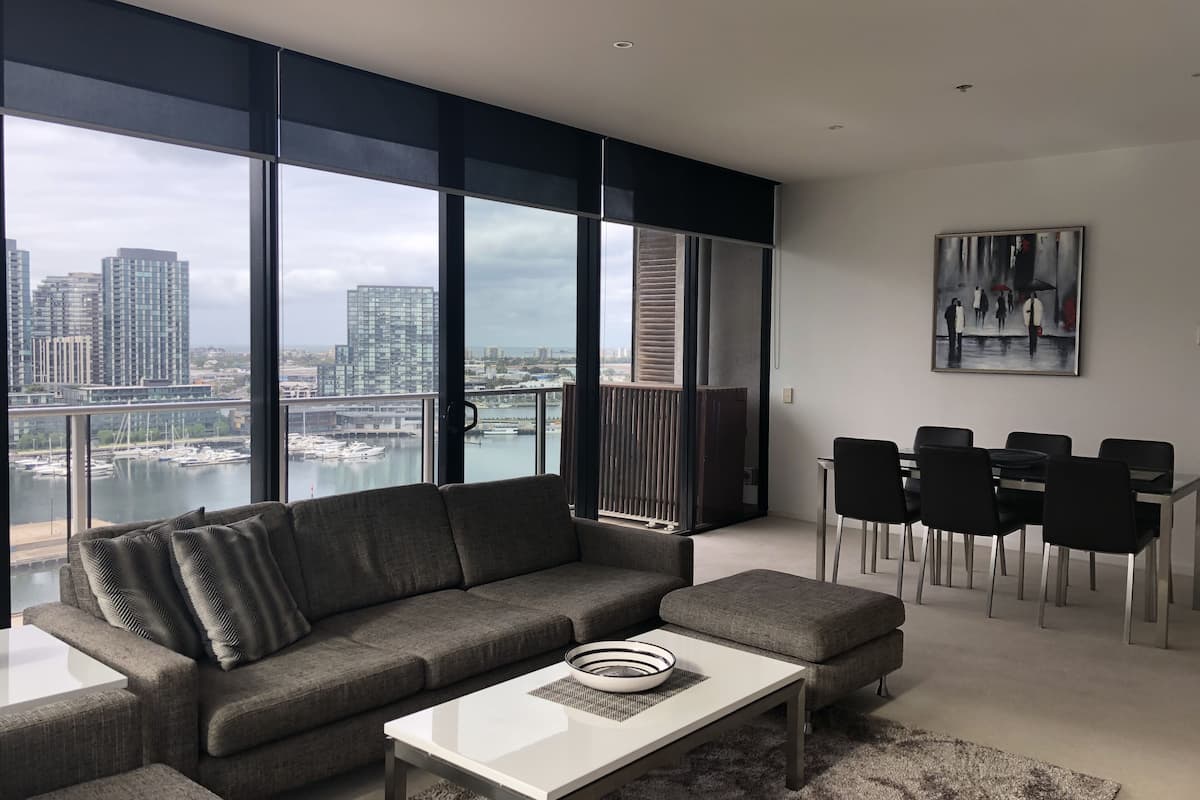 Una sala de estar moderna con un sofá seccional gris, una mesa de centro blanca y un comedor con sillas negras. Amplias ventanas ofrecen vistas a un paisaje urbano y un puerto deportivo.