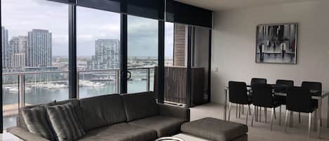 Un salon moderne avec un canapé d’angle gris, une table basse blanche et un coin repas avec des chaises noires. De grandes fenêtres offrent une vue sur le paysage urbain et le port de plaisance.