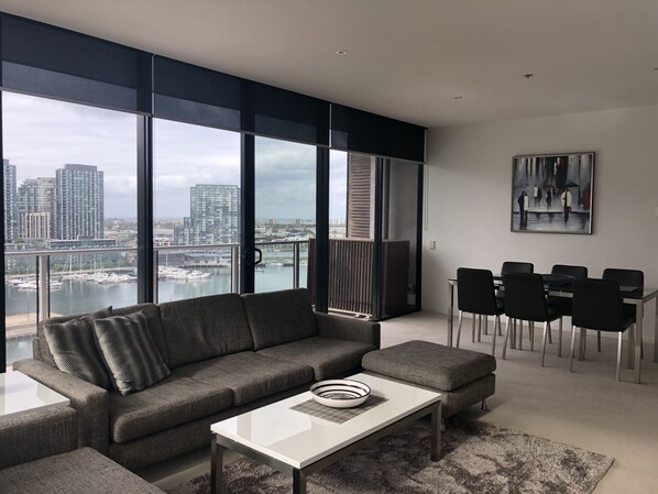 A modern living room with a gray sectional sofa, a white coffee table, and a dining area with black chairs. Large windows offer a view of a cityscape and marina. - Accent Accommodation at Docklands Melbourne (Docklands)