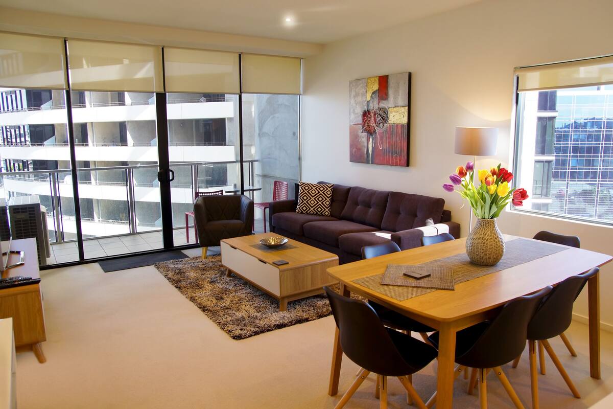 Una sala de estar moderna con sofá, mesa de centro y mesa de comedor. Hay un ventanal con vista a un edificio.