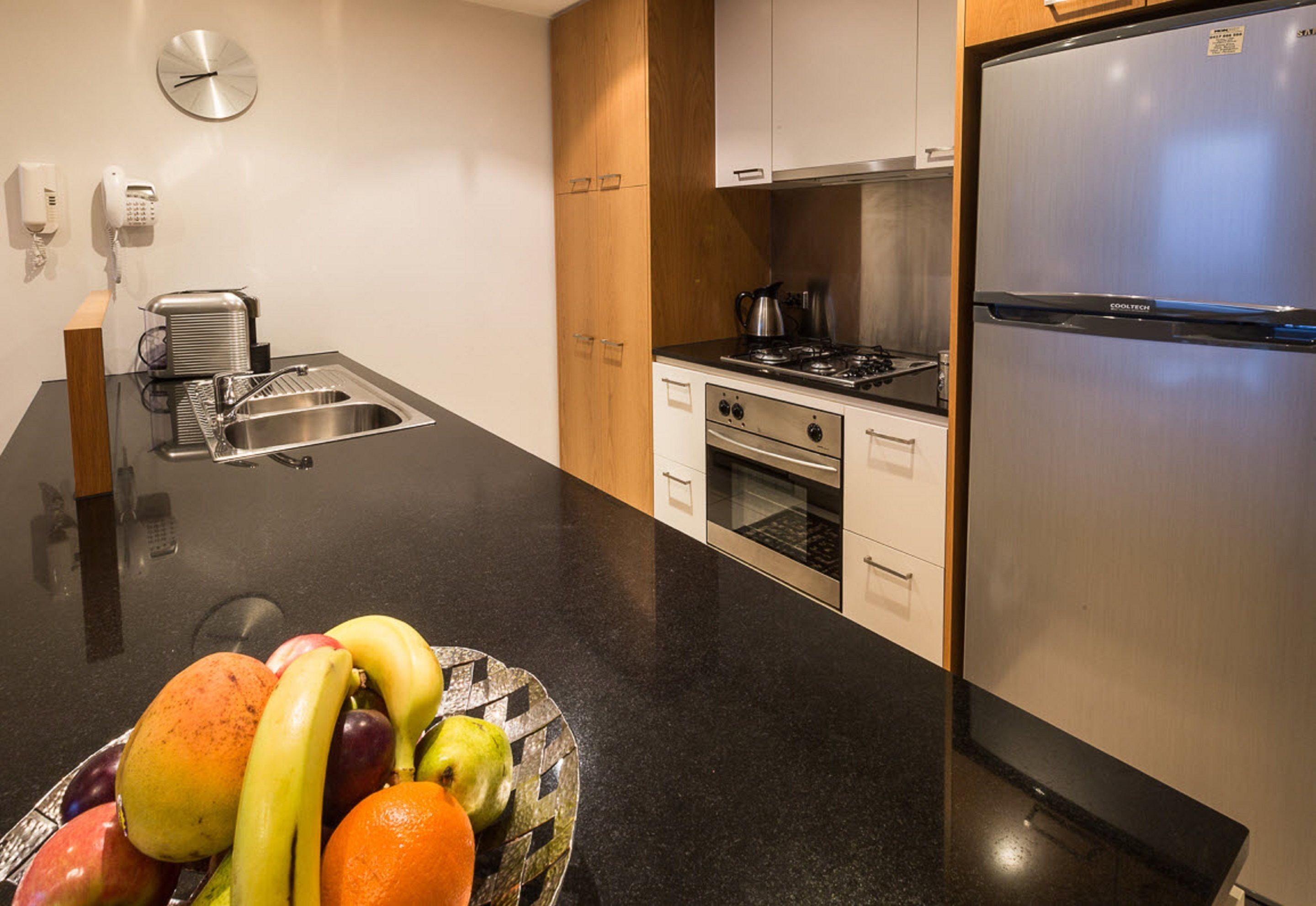 A modern kitchen with a stainless steel refrigerator, a stove, and a sink. There is a fruit bowl on the counter and a clock on the wall.
