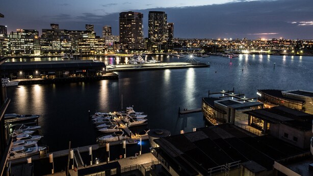 A marina with boats docked, a city skyline in the background, and buildings with illuminated windows.