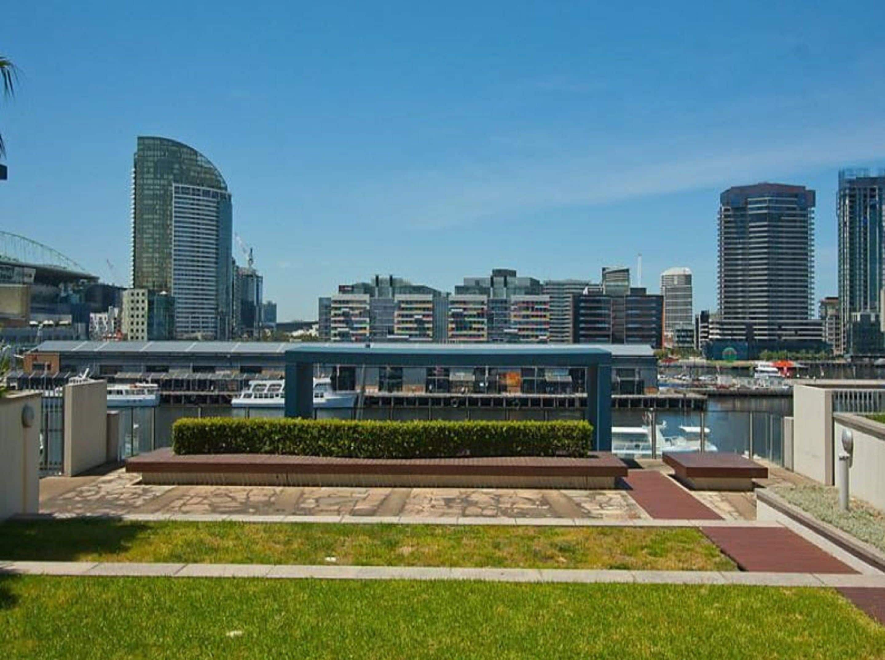 A modern urban waterfront area with a view of high-rise buildings, a marina, and a pedestrian walkway with a landscaped area.