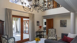 A spacious living room with a chandelier, a grey sofa, patterned chairs, and a white coffee table with a vase of flowers. - Villa Tuscana By Mantis (Gqeberha (Port Elizabeth))