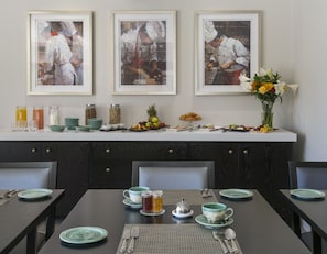 Un comedor con mostrador blanco que exhibe alimentos para el desayuno, tres obras de arte enmarcadas encima del mostrador, y un jarrón con flores a la derecha.