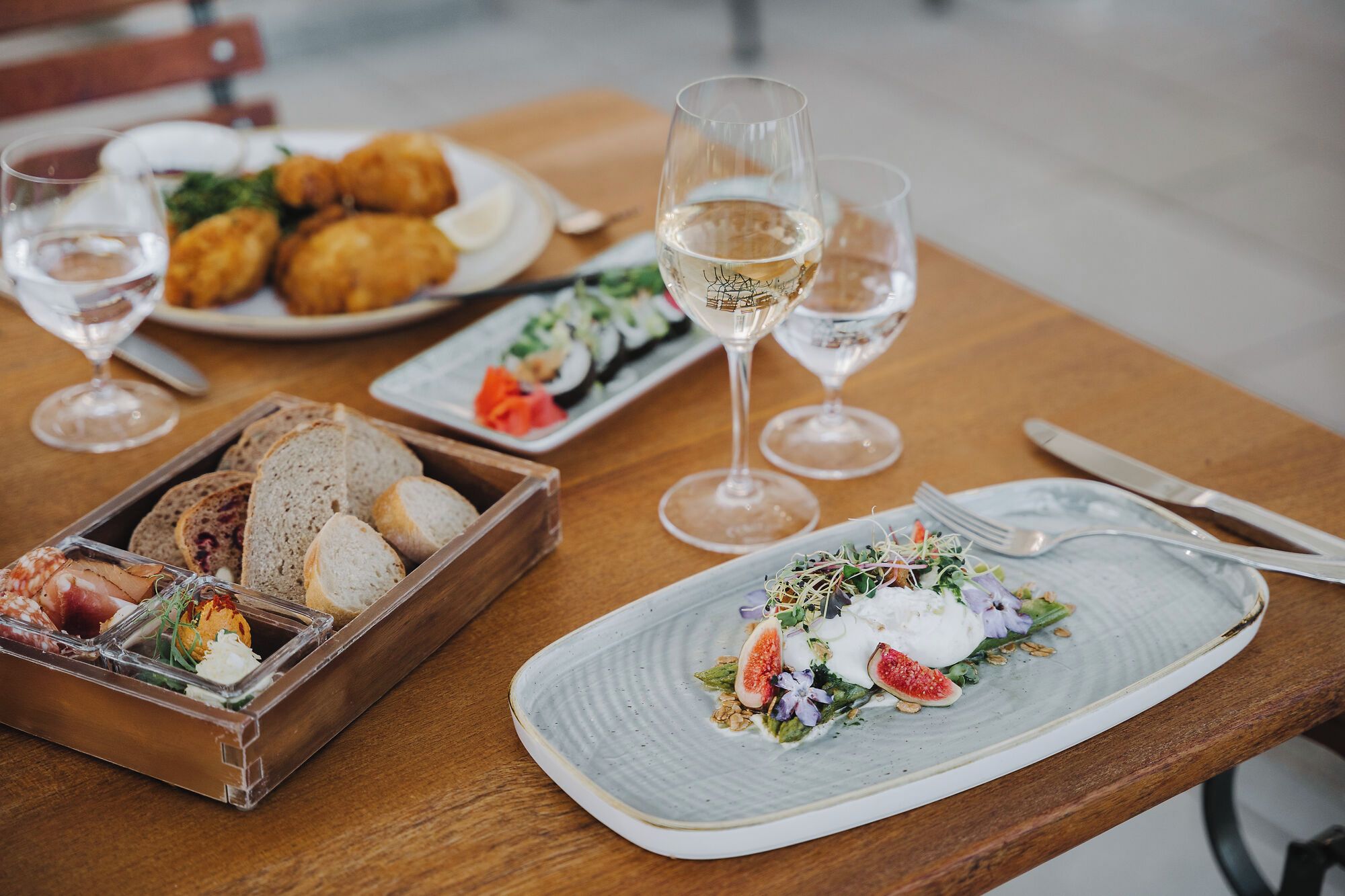Uma mesa de jantar com diversos pratos, copos de vinho branco e uma cesta de pão.