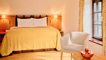 Ein Hotelzimmer mit einem Bett, Nachttischlampen, einem Sessel, einem kleinen Tisch mit Obstschale und einem Fenster mit Vorhängen.