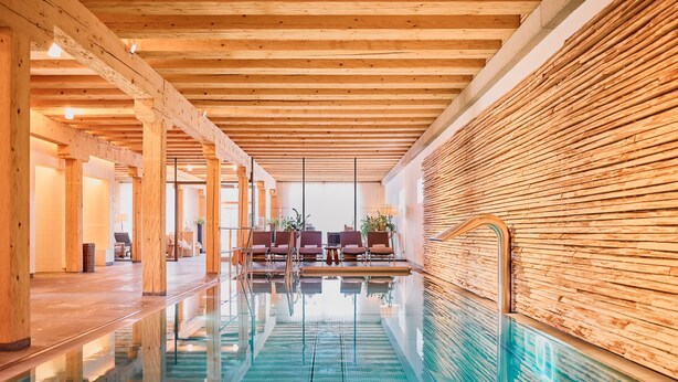 Une vaste piscine intérieure dotée de poutres en bois et d’un sol carrelé, entourée d’espaces de détente et d’un mur en bois.